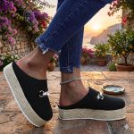 Women's black canvas platform mule espadrilles featuring a textured jute rope sole and a sparkling gold chain buckle, set against a Mediterranean coastal patio backdrop.