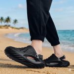 Lifestyle shot featuring a model's feet wearing black Piki closed-toe recovery slides, paired with black athletic ankle socks and grey jogger pants.